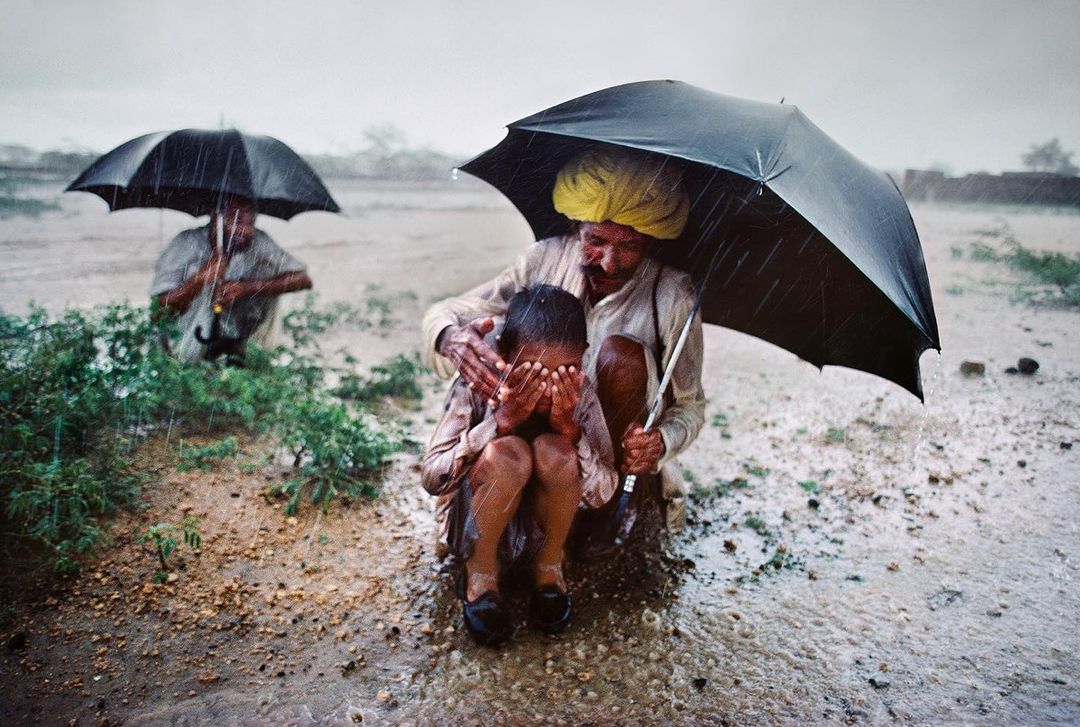 A child huddled under an umbrella held by an older adult, both kneeling in a field during a downpour, with another figure in the background.