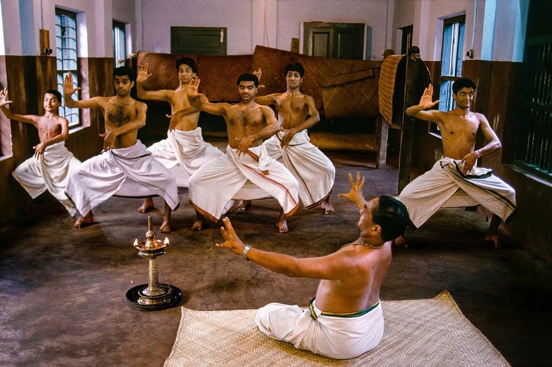 Group of Indian men in traditional white dhotis performing a dance indoors with a brass oil lamp in the foreground.
