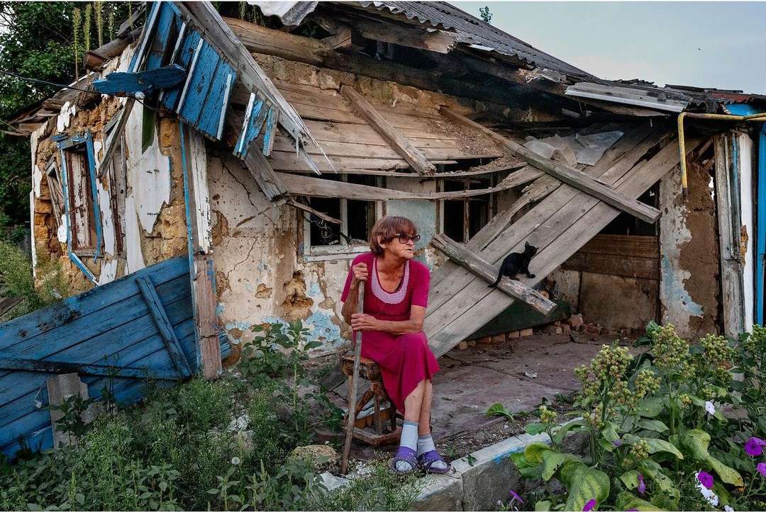 Elderly woman sits outside a damaged house, next to overgrown vegetation, with a black cat on the broken roof and debris visible.