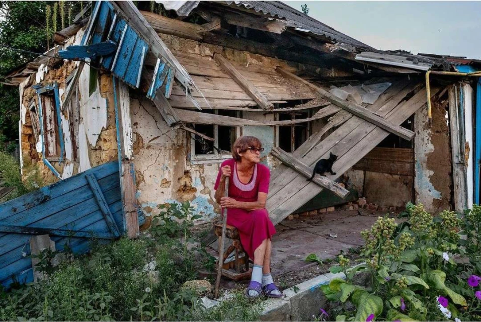 Elderly woman sits outside a damaged house, next to overgrown vegetation, with a black cat on the broken roof and debris visible.