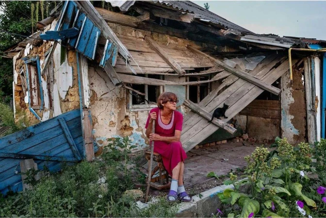 Elderly woman sits outside a damaged house, next to overgrown vegetation, with a black cat on the broken roof and debris visible.