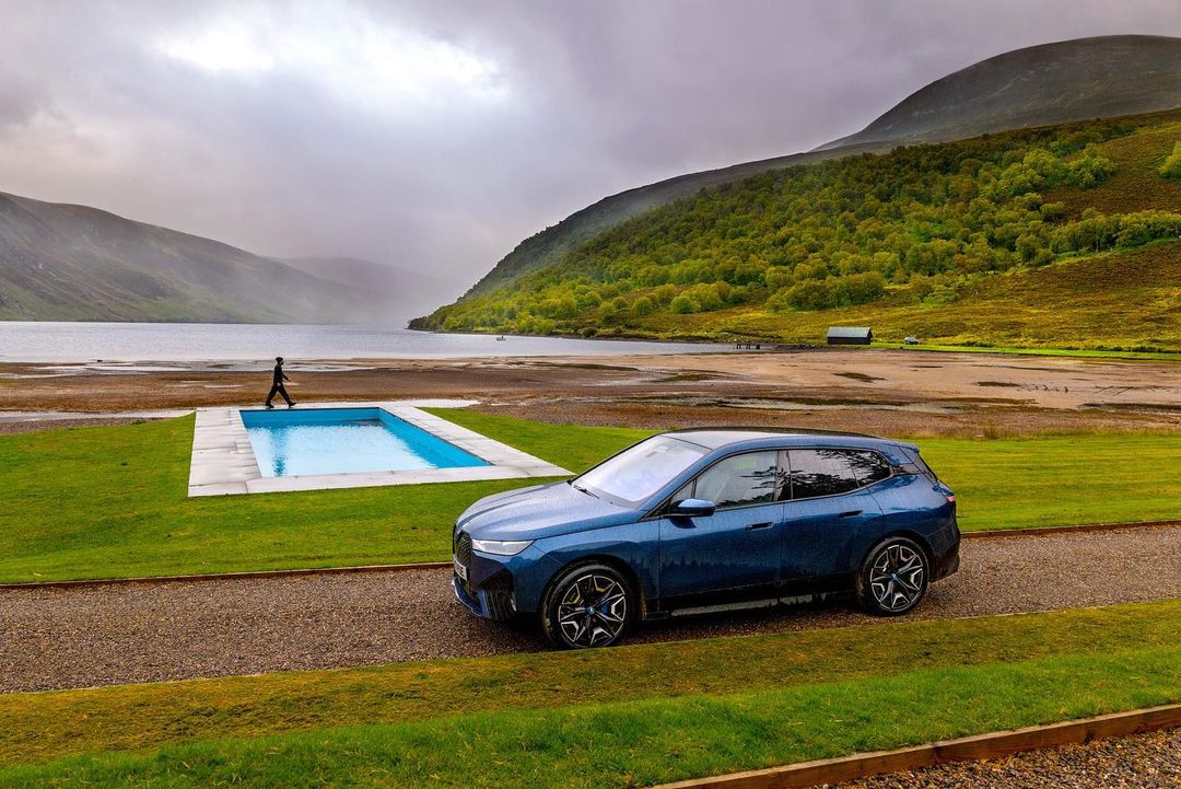 Blue SUV parked on a gravel road with a swimming pool and lake in the background, surrounded by green hills and a cloudy sky.