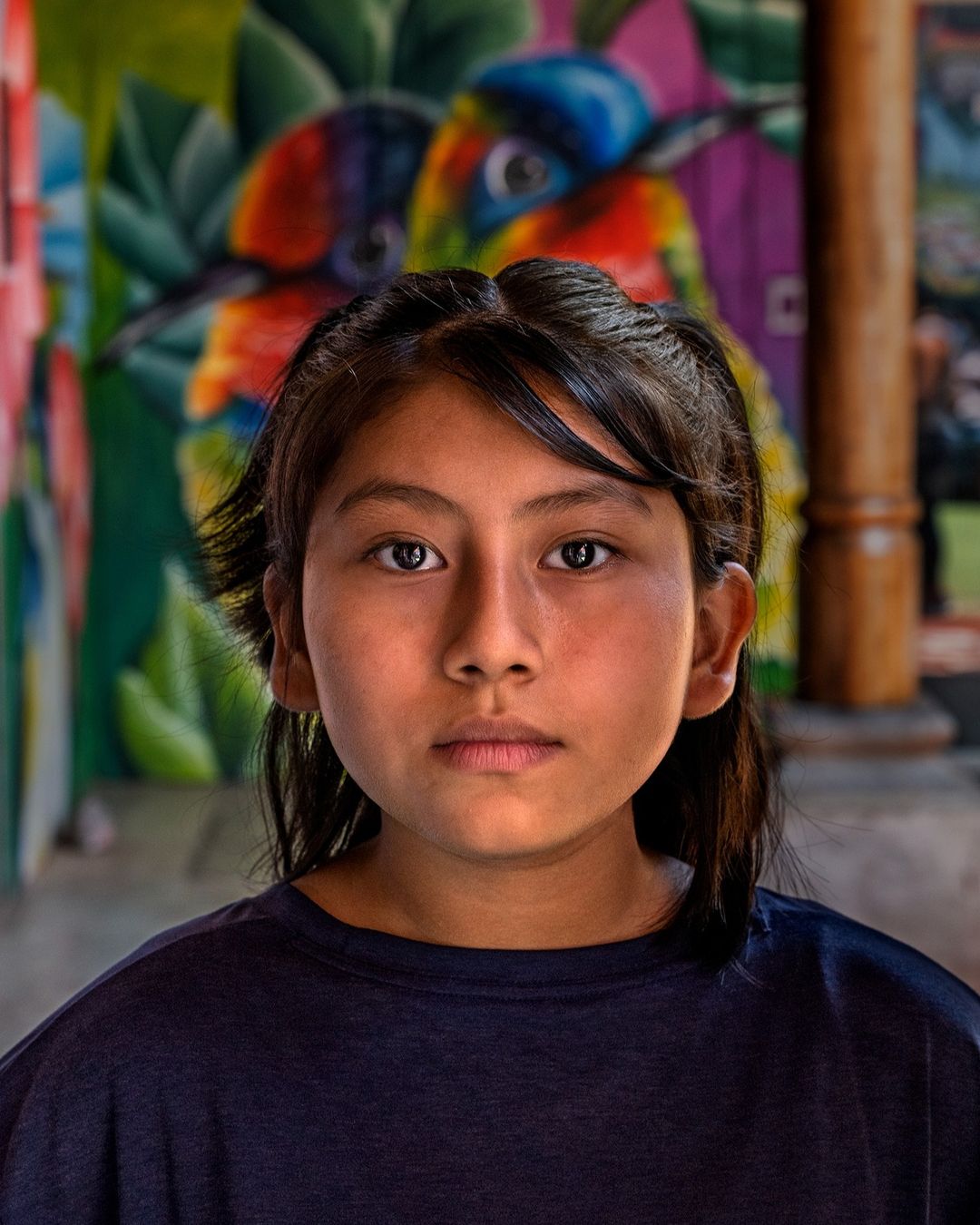 Close-up portrait of a young indigenous girl with a serious expression, set against a vibrant mural background in a colorful indoor space.