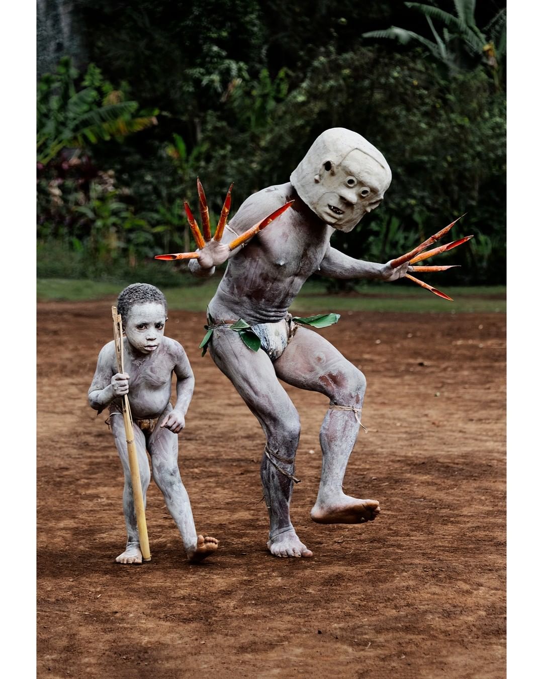 Two people are performing in an outdoor environment; the person in the background is wearing a mask and claw-like finger extensions.