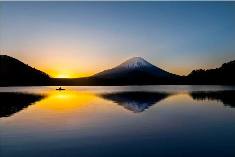 Scenic landscape view of Mount Fuji and its reflection on a lake, with a silhouette of a person in a boat, during a golden sunset.