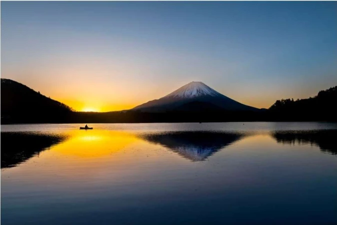 Scenic landscape view of Mount Fuji and its reflection on a lake, with a silhouette of a person in a boat, during a golden sunset.