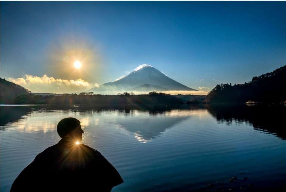 Person silhouetted by the sun, gazing across a calm lake towards a majestic mountain partially obscured by clouds.