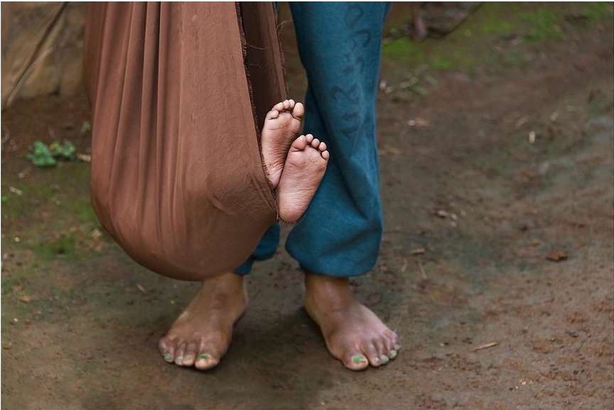 Low-angle view of a child's feet protruding from a brown sling, with an adult's legs and feet standing behind, outdoors on the ground.
