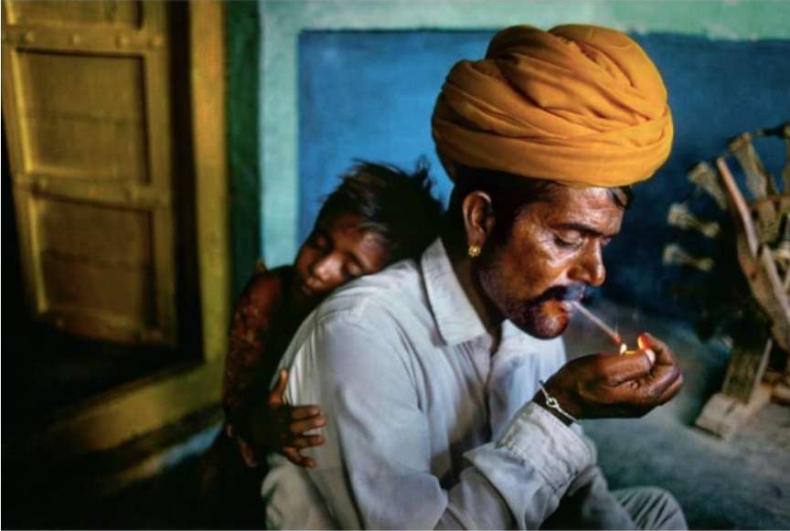 Man in turban lights cigarette as child rests on his shoulder indoors, displaying warmth and connection in a candid portrait.