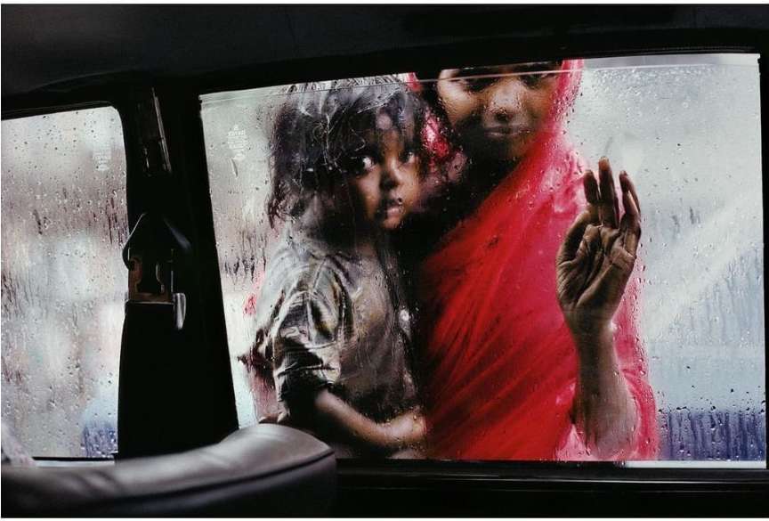 Rainy day portrait captures an adult and child in a taxi, separated from the blurred city backdrop by a wet, streaked car window.