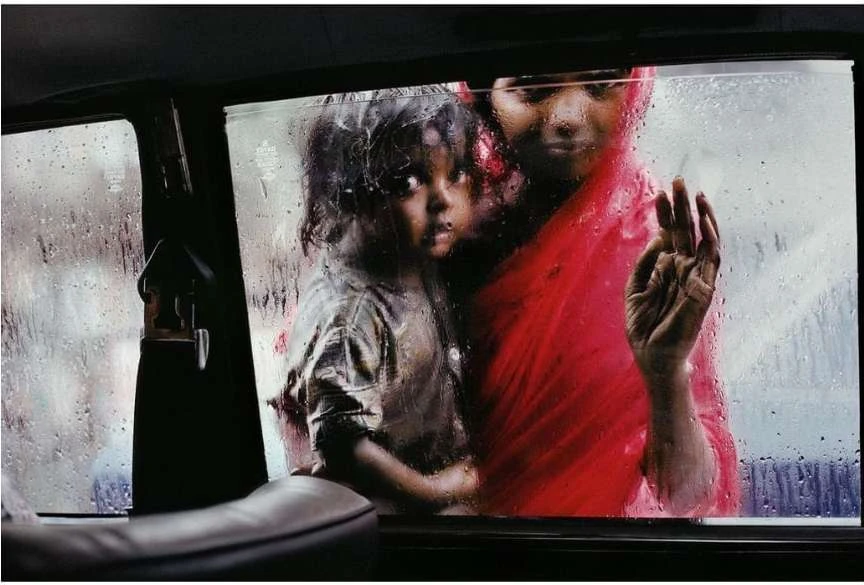 Rainy day portrait captures an adult and child in a taxi, separated from the blurred city backdrop by a wet, streaked car window.