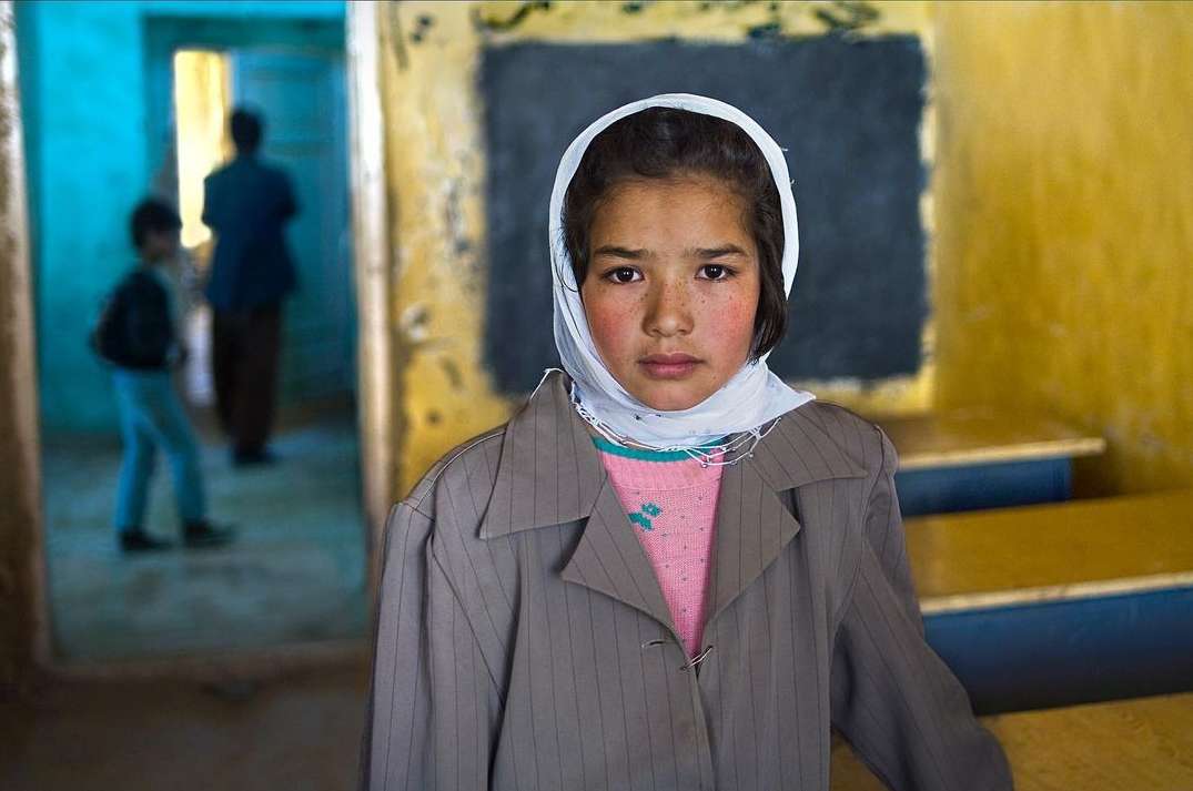 Portrait of a young girl wearing a headscarf and jacket, standing indoors with blurred figures in the background, creating a sense of place.