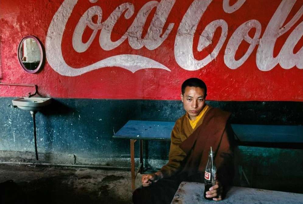 Monk sits at a table holding Coca-Cola bottle in an indoor setting with a large Coca-Cola sign on the red and blue wall.