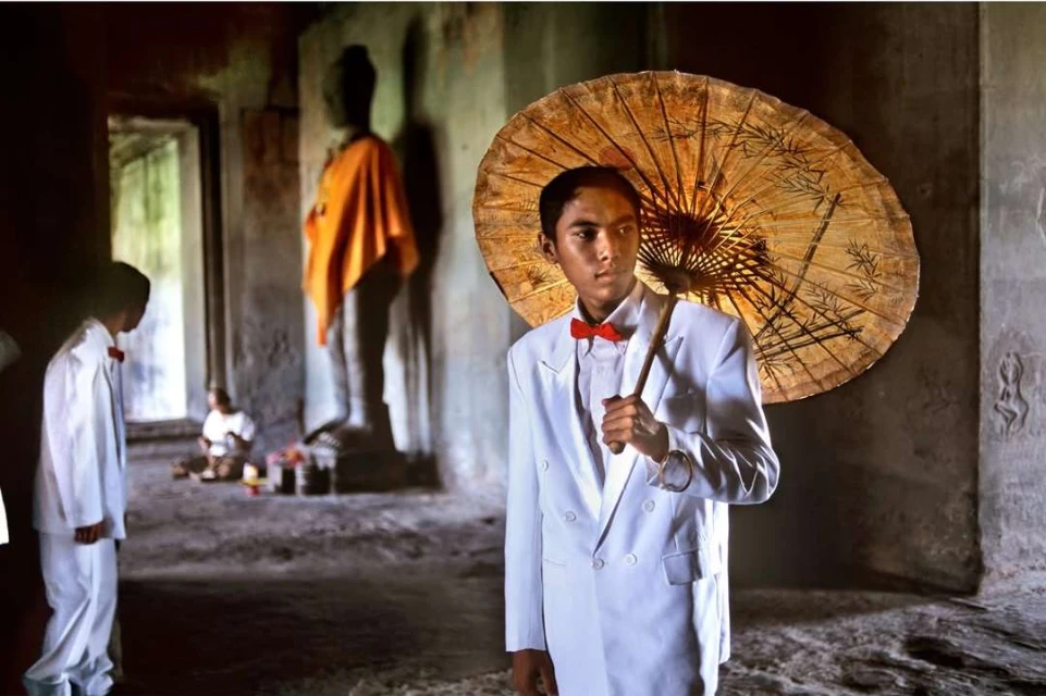 Young adult poses in a white suit and red bowtie, holding an umbrella with temple ruins and a Buddha statue in the background.