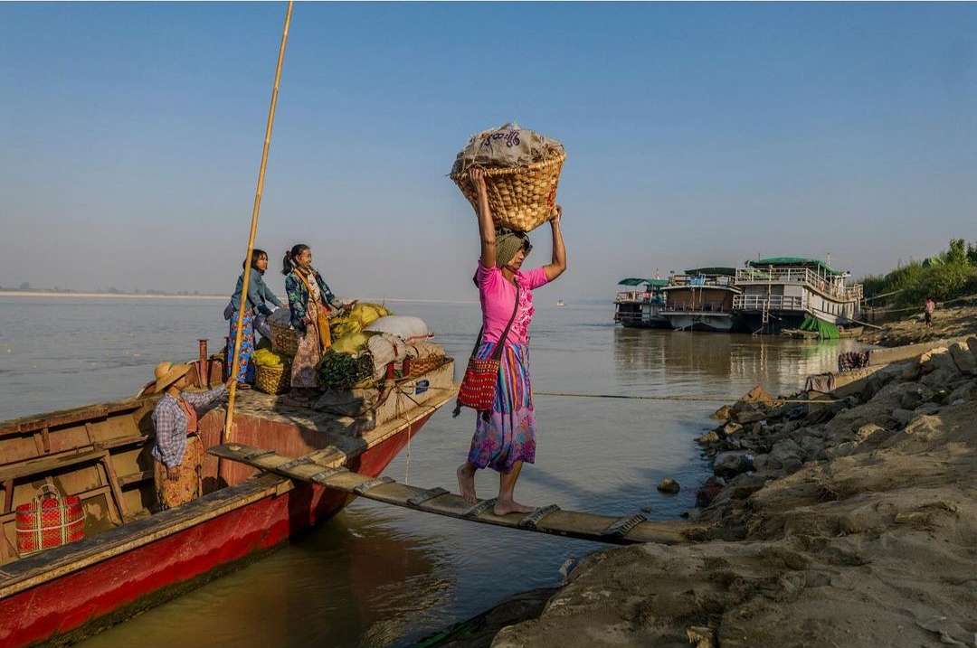 Woman balances a basket on her head while crossing a wooden plank from a boat to a riverbank, with other people on the boat.