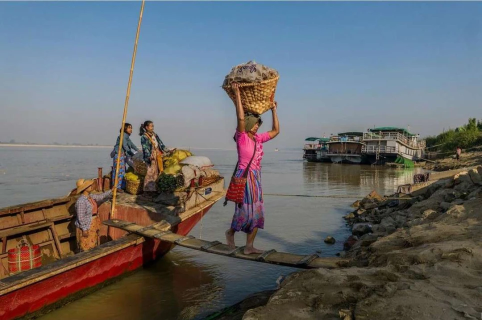 Woman balances a basket on her head while crossing a wooden plank from a boat to a riverbank, with other people on the boat.