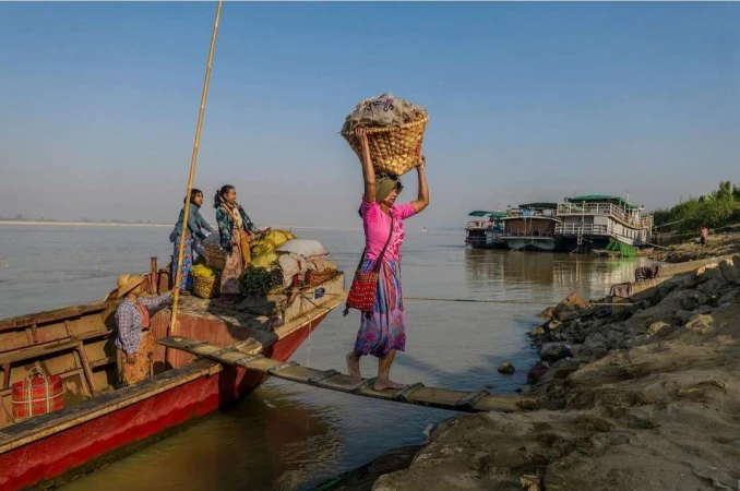 Woman balances a basket on her head while crossing a wooden plank from a boat to a riverbank, with other people on the boat.