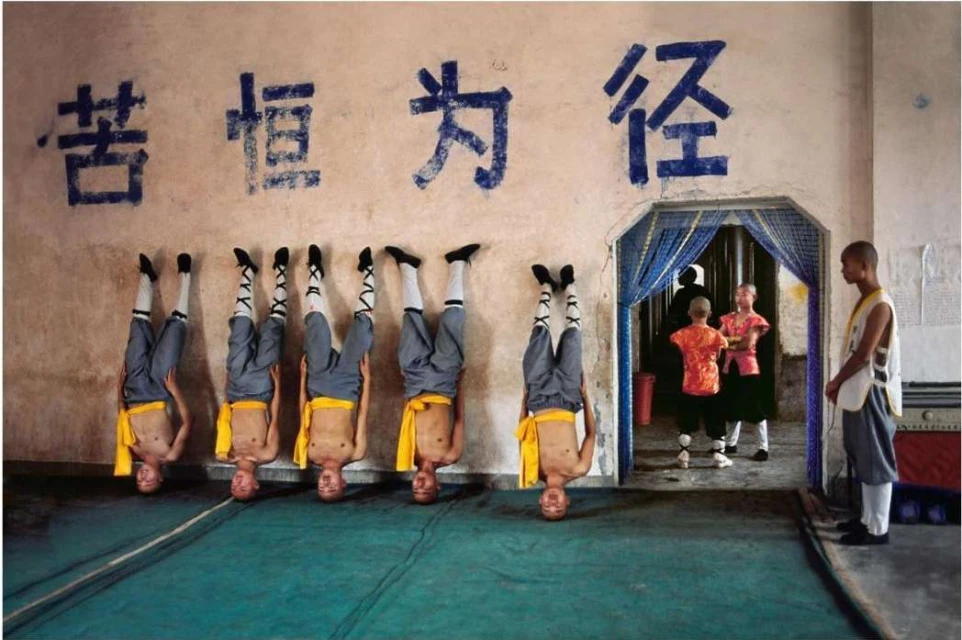 Group of Shaolin monks demonstrating headstand against a wall with Chinese writing in an indoor space with people in the background.