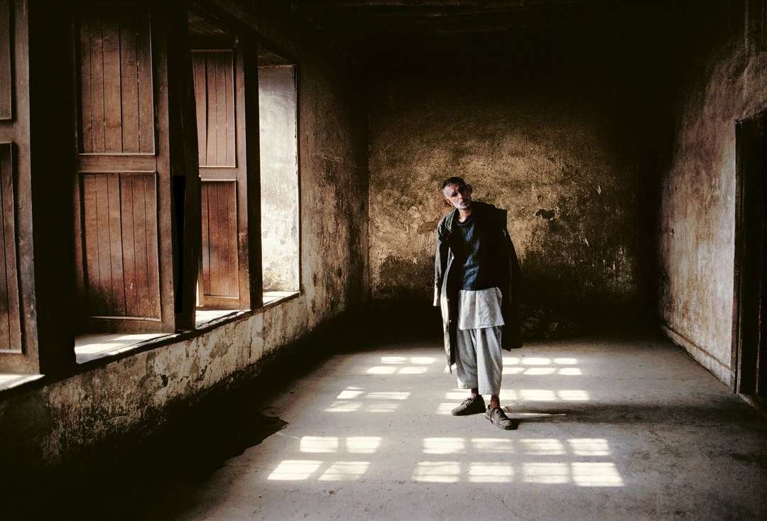 Indoor shot of a thin man in a jacket standing in a weathered room with light streaming through window shutters, creating shadows.