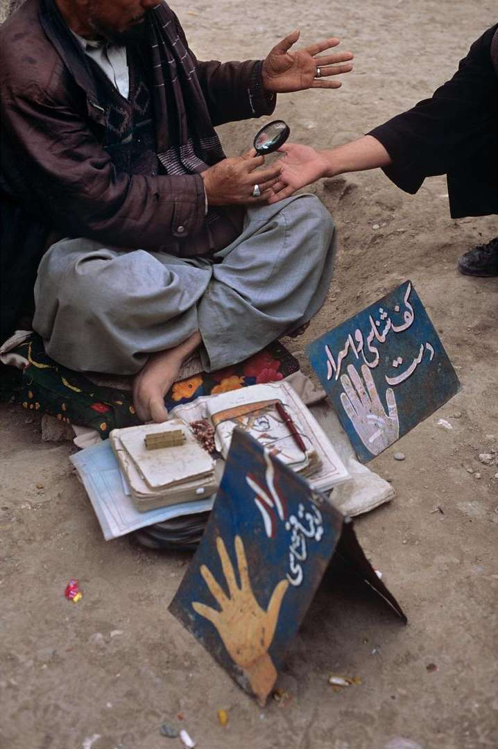 An elderly man in casual attire sits outdoors, examining a young person's hand with a magnifying glass, surrounded by books and signage.