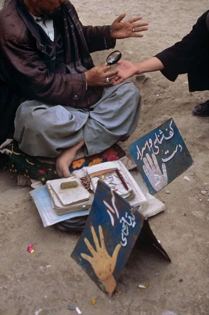 An elderly man in casual attire sits outdoors, examining a young person's hand with a magnifying glass, surrounded by books and signage.
