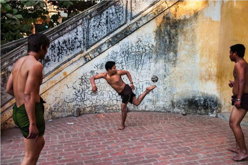 Three shirtless young adult men play a game with a soccer ball on a brick surface with a wall and stairs behind them.