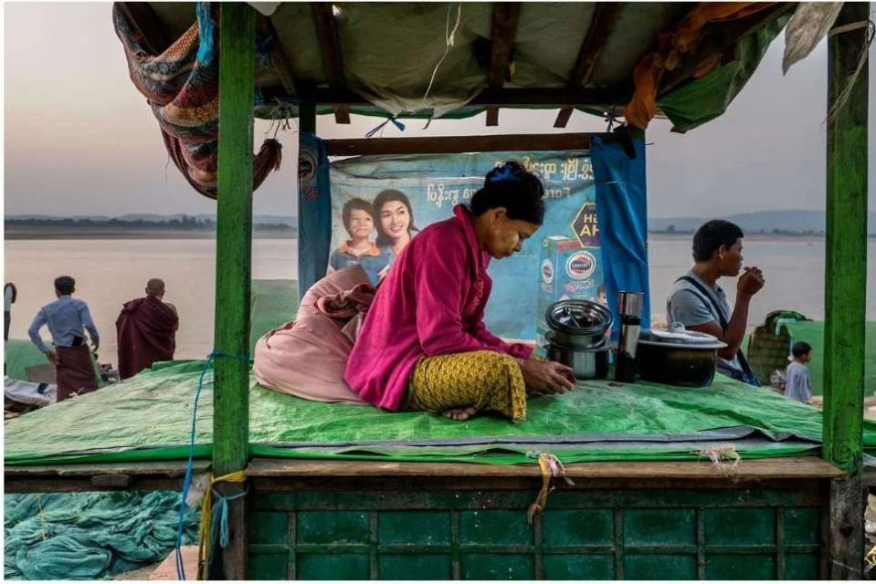Outdoor scene features an older Asian woman preparing food at a roadside stall by a river with other people in the background.