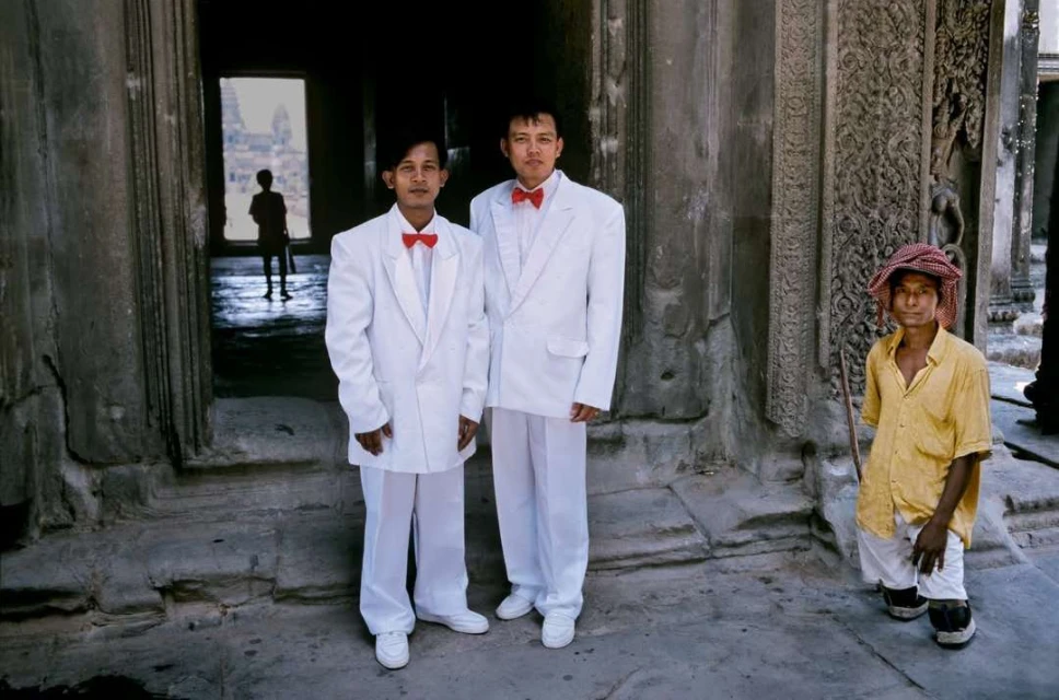 Two men in white suits with red bow ties stand in front of ancient stone architecture, with a third man to the side.