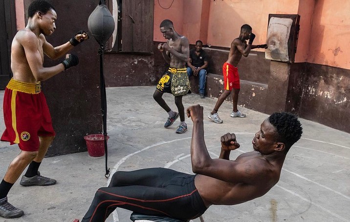 Several shirtless men train in an indoor boxing gym. Some practice punches, while one does sit-ups in the foreground.