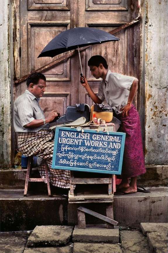 Two men at a street-side writing service, one seated at a typewriter, the other holding an umbrella, in front of a rustic door.