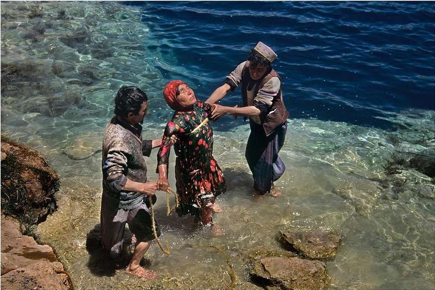 Two men assisting an older woman in the water near a rocky shore. The woman is wearing a colorful dress and headscarf.