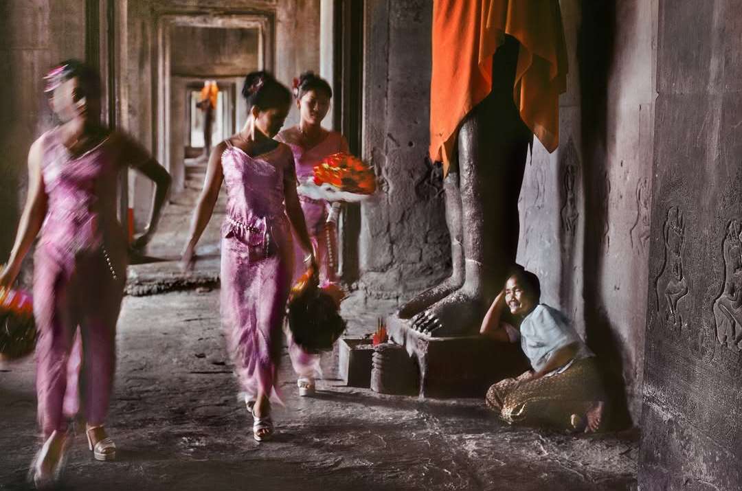 Three women in pink dresses walk in procession inside an ancient stone temple with columns, while a fourth woman sits on the floor.