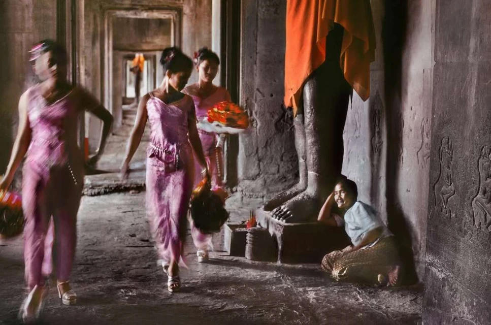 Three women in pink dresses walk in procession inside an ancient stone temple with columns, while a fourth woman sits on the floor.