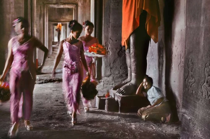 Three women in pink dresses walk in procession inside an ancient stone temple with columns, while a fourth woman sits on the floor.