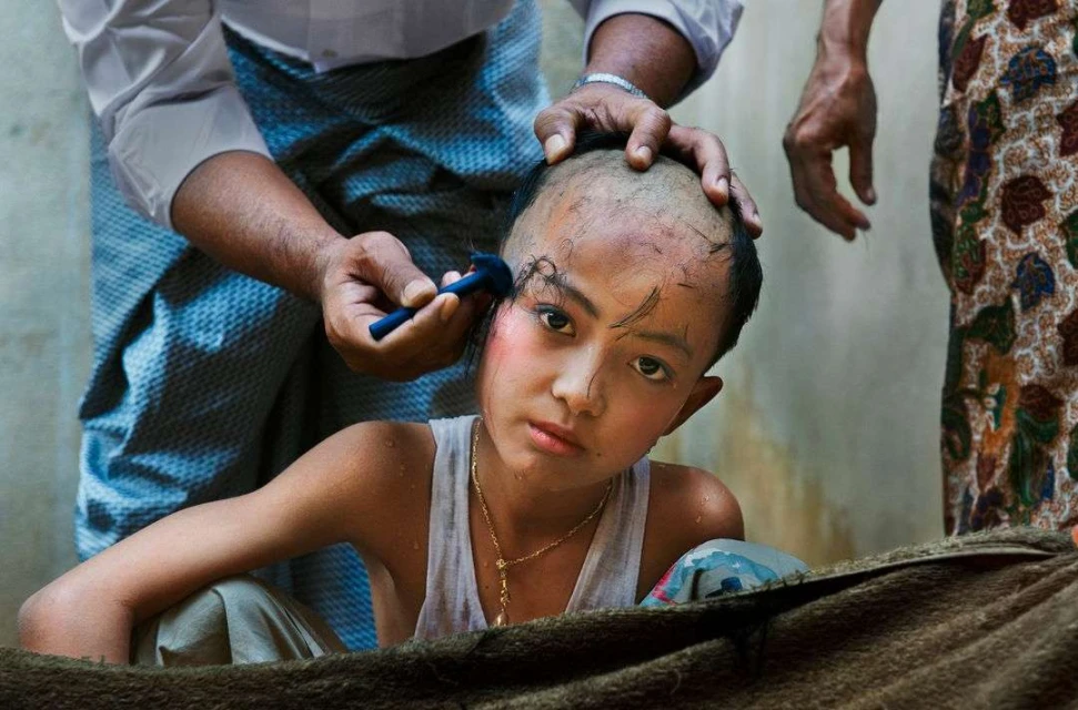 Child receiving a ceremonial haircut, with an adult male using a razor to cut remaining hair, as part of a cultural or religious tradition.
