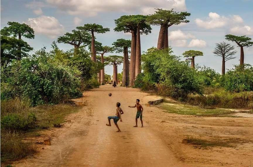 Two young boys play soccer on a dirt road lined with baobab trees in a rural setting under a partly cloudy sky, outdoors.