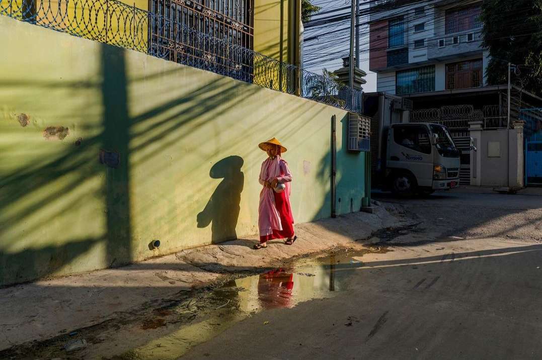 Adult woman in a pink robe and straw hat walks along an urban street, her shadow cast on a pale-yellow wall near a small truck.