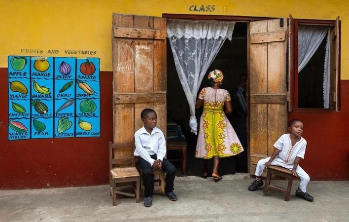 Children seated outside a colorful school building with painted fruit illustrations on the yellow and red wall.