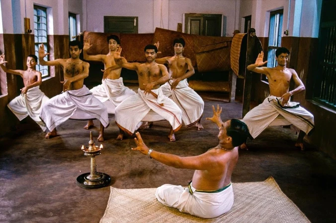 Group of six South Asian male dancers practicing traditional poses instructed by a seated teacher by a small lamp.