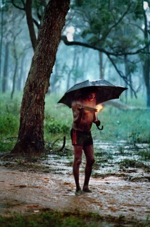 Man holding an open umbrella and walking barefoot through a muddy flooded forest path during heavy rain.