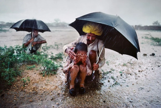 An older man holding a black umbrella over a child crouching in the rain, with a second figure under an umbrella in the background.