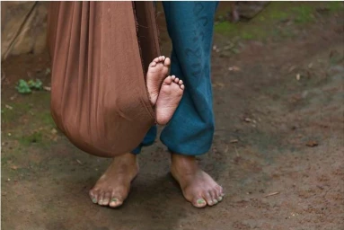 Close-up portrait of a person standing next to a makeshift fabric hammock containing an infant with visible feet.