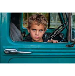 Young boy with curly blonde hair and striking blue eyes looking out from the open window of a teal truck