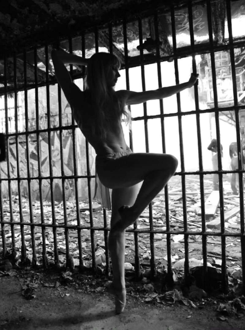 Black and white photo of a dancer in pointe shoes striking a pose behind metal bars, inside a decayed, derelict structure with a concrete floor.