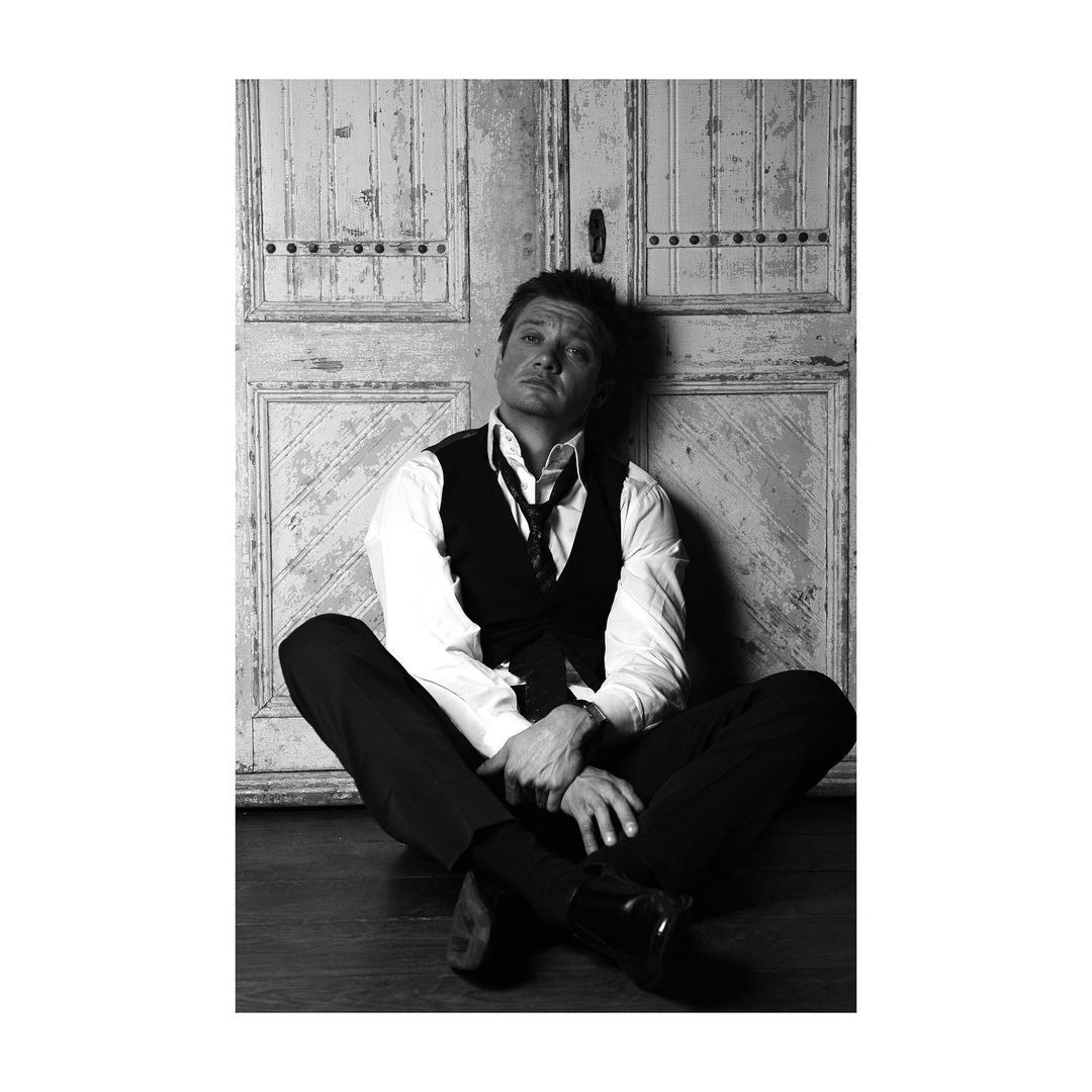 Monochrome portrait of a man in a vest and tie sitting in front of a weathered wooden door. Black and white fashion style.