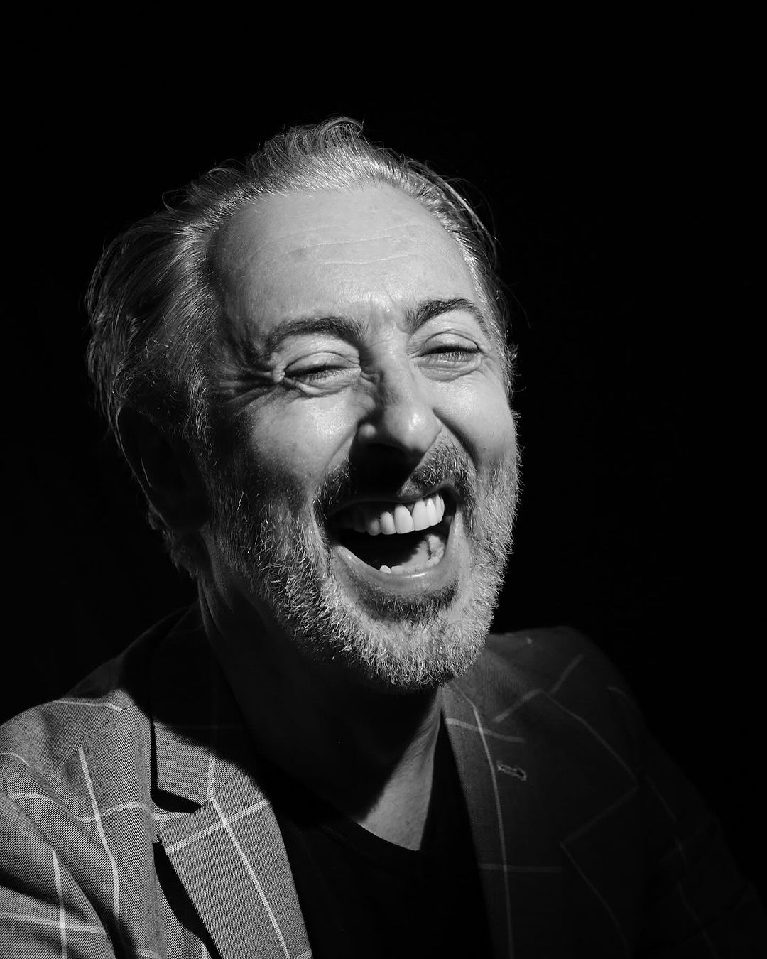 Monochrome portrait of a middle-aged man laughing heartily while wearing a plaid blazer against a dark background, head and shoulders.
