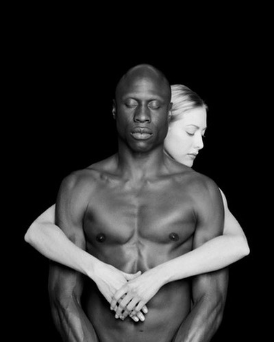 Striking black and white portrait of a shirtless African-American man embraced from behind by a fair-skinned Caucasian woman.