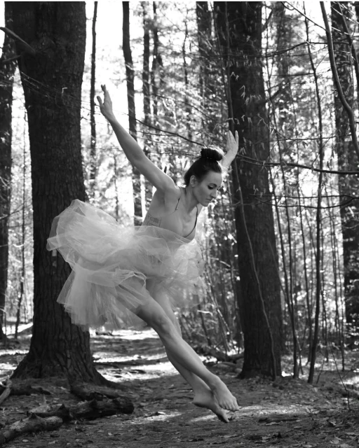 Ballerina in white tutu leaps barefoot through a forest clearing, sunlight filtering through the trees, black and white.