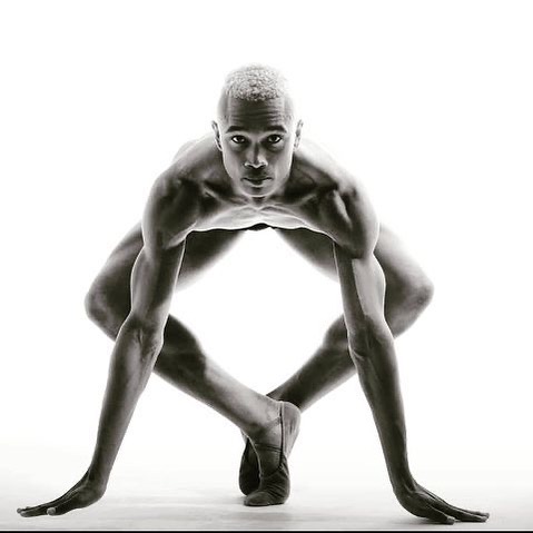 Artistic black and white studio portrait of a flexible adult male ballet dancer crouching with hands on the floor looking at the camera.
