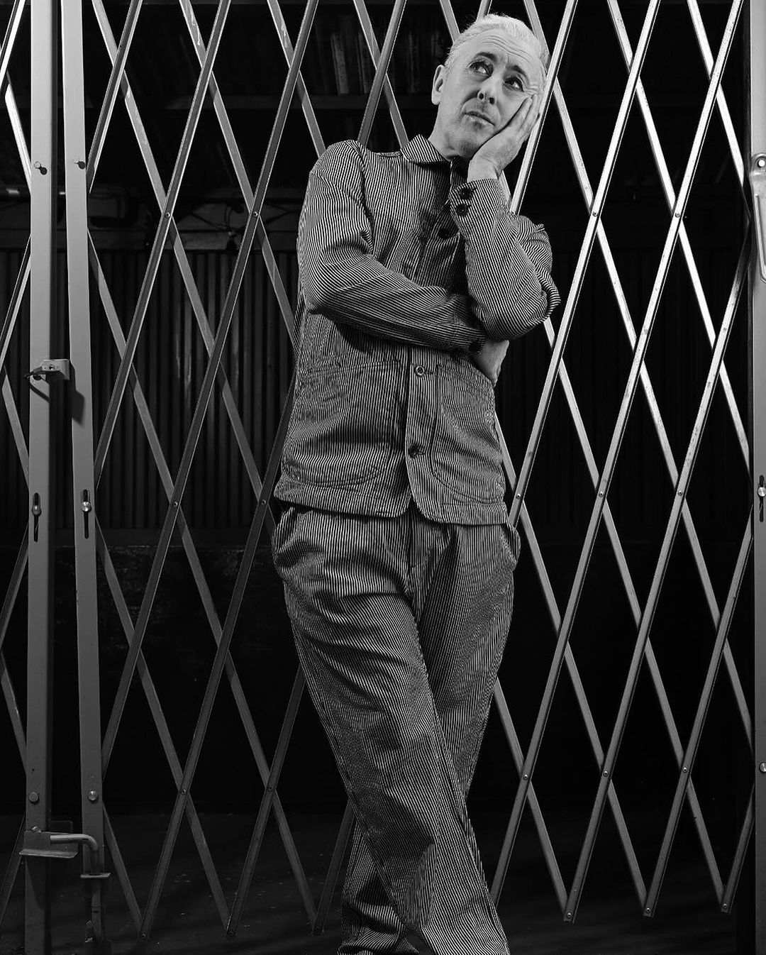Monochrome portrait of a pensive adult male with silver hair leaning against a metal gate wearing striped jacket and trousers.